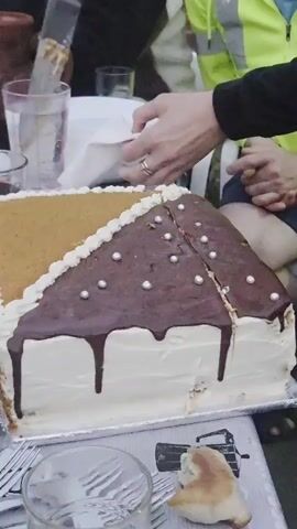 A cake is being cut on a table. The cake is rectangular and decorated with white frosting, a chocolate glaze, and a honey-flavored top layer. It is adorned with small silver pearl decorations and chocolate drizzles. Two people are visible; one person is wearing a green high-visibility vest and is partially cut out of the frame, while another person, whose hands are visible, is cutting the cake with a knife. There are plates and glasses on the table, as well as a pitcher in the background. The environment appears to be outdoors, possibly a gathering or celebration.