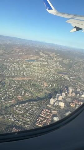 The video shows an aerial view of a city with a clear blue sky. An airplane wing is visible in the upper right corner. The city below is densely populated with residential buildings and a commercial district. A golf course is also visible, along with bodies of water. The video is shot from inside an airplane, looking out the window. The time stamp indicates the video is from 00:00 to 00:04.