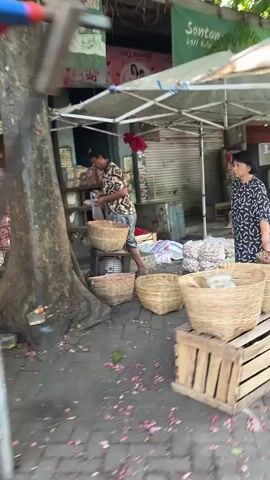 The video shows a bustling outdoor market scene. In the foreground, several large woven baskets are filled with what appear to be onions or shallots, arranged on wooden crates. The baskets are positioned on the left and right sides of the frame. Two individuals are visible in the middle ground. On the left, a person wearing a patterned shirt is interacting with a large pile of produce. On the right, a person wearing a black and white patterned top is near a basket of produce. In the background, more produce, possibly onions, is stacked in large quantities, and stalls with awnings suggest a marketplace setting. The lighting indicates it is daytime, and the overall atmosphere is lively.