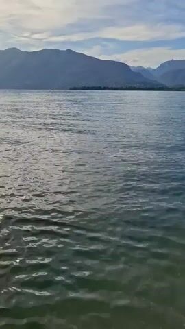 A drone shot moves forward over a body of water. In the background, mountains rise up, covered in greenery and dotted with buildings. The sky above is partly cloudy, with the sun casting a soft light on the landscape. The water is rippling gently, with its surface reflecting the sky and mountains. The scene is serene and peaceful.