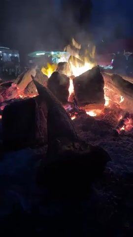 A bonfire burns at night, with large logs of wood stacked and actively flaming. The fire casts a warm, orange glow on the surrounding snow. In the background, out of focus, are what appear to be food vendors and people gathered around the fire, silhouetted against the dim light of the evening sky. The scene suggests a festive outdoor gathering or event.