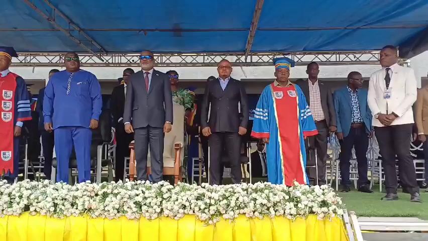 A group of people, mostly men, stand on a platform decorated with white flowers and a yellow cloth. There are 11 people visible, some wearing academic robes and hats, others in suits or traditional clothing. A blue tarp forms a canopy above them. After an announcement, the people bow in unison. The setting appears to be an outdoor event, possibly a graduation or ceremony.