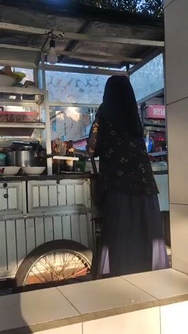 A street food cart is seen from the side. The cart has shelves stacked with various food containers and utensils. A person wearing a dark headscarf and a patterned shirt is standing behind the cart, appearing to be preparing food. In the background, a street scene unfolds with motorcycles parked, buildings, and power lines overhead. The lighting suggests it is either early morning or late afternoon.