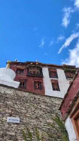 A low-angle, vertical video captures a stone wall leading up to a multi-story building with distinct red and white facades. A white stupa with a gold spire is visible on the left. The building has multiple windows with wooden frames and shutters, some open, some closed. The sky above is a vibrant blue with scattered white clouds. A signpost on the wall reads "WAY TO MONASTERY" with an arrow. Green foliage grows at the base of the wall. The overall scene suggests a traditional architectural style in a mountainous or high-altitude environment.