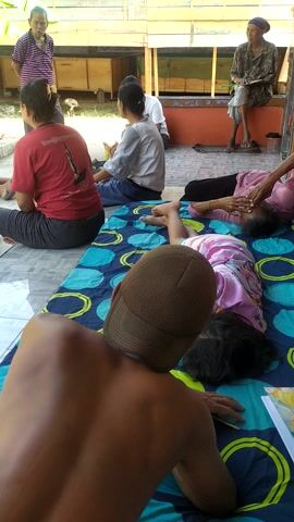 This media file captures an outdoor gathering of several people, likely in Desa Penggalang, Kecamatan Adipala, Indonesia, given the visual cues. The scene unfolds on a tiled patio with a backdrop of banana trees and a wooden structure, suggesting a rural or semi-rural setting. The time of day appears to be daytime, indicated by the bright sunlight casting shadows.

In the foreground, a person wearing a brown baseball cap is partially visible from the back, lying on a blue and green patterned mat. Next to them on the mat is a child in a pink shirt. Further back, a group of women are seated or resting on mats and the ground. One woman in a red t-shirt is facing away from the camera, while another in a white long-sleeved shirt is also seated. An older woman in a striped shirt stands further back, and another older woman in traditional clothing sits on a raised bench, holding a book or document. A few pairs of flip-flops are scattered on the ground, suggesting a casual atmosphere.

The overall impression is one of a relaxed social gathering, possibly a family or community event. The people appear to be conversing and interacting, though the specific activity is not definitively clear. Some individuals seem to be receiving attention, with one woman appearing to massage another's head. The expressions on the faces of those visible suggest a calm and possibly pleasant mood.

Visible text includes "DIRGAHAYU" and possibly "TERUS" on a banner in the background, hinting at a celebratory occasion or a local event. The presence of what appears to be a government ministry name on a sign also points to a community-related activity. The weather appears to be warm and sunny, suitable for an outdoor gathering.
