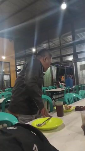 A man in a black jacket is standing by a table at what appears to be an indoor restaurant. He is looking down at the table and has his hands near a plate. In front of him is a green plate, a spoon, and a glass of dark liquid. Several green plastic chairs are visible, along with tables covered in white tablecloths. In the background, there are more tables and chairs, as well as a serving counter with people working behind it. The lighting appears to be artificial, with overhead lights visible. The overall impression is of a casual dining establishment.