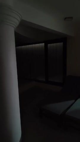 This media file appears to be a short video clip or a series of still images captured at night. The initial scenes show an indoor setting with a large white pillar on the left and dark lounge chairs in the foreground. In the background, there are large glass doors with vertical blinds, through which faint light is visible. The overall ambiance is dim and appears to be a spa, lounge, or hotel room.

The scene then transitions to an outdoor view from a window or balcony at night. A full moon is visible in the dark sky. Below, there is a silhouette of buildings with lights on in some windows. The view overlooks what appears to be a landscape with trees and possibly a body of water or a dark expanse. Streetlights or decorative lights are also visible, casting a warm glow. The overall impression is one of a quiet, serene night, possibly in a resort or a residential area.

The text "00:00" is visible at the beginning of the clip, indicating a timestamp. The spoken words "Красиво так" (Beautiful like this) and "подсвечиваются окошки" (the windows are lit up) are heard, suggesting the speaker is appreciating the view. The words "балкон" (balcony) are also spoken, reinforcing the interpretation of an outdoor view from a higher vantage point. The location context provided, Phu Thuy Ward, Vietnam, adds to the understanding of the setting. The time of day is clearly night, and the weather appears clear enough for the moon to be visible. The emotions conveyed are of appreciation and peacefulness.