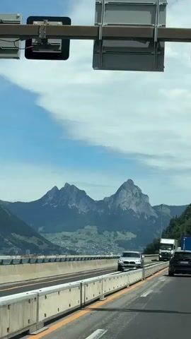 A landscape view from a moving vehicle on a highway shows a bright blue sky with scattered clouds. In the distance, majestic mountains with snow-capped peaks dominate the horizon, with green hills and a body of water visible below them. The highway is bordered by concrete barriers and has multiple lanes of traffic. Several vehicles, including a white truck, a silver car, and a black car, are traveling on the highway. The scene is illuminated by daylight, suggesting it is daytime.