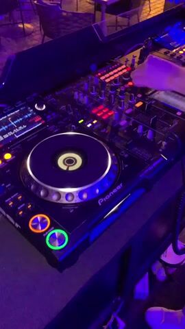 A close-up shot shows a DJ's hands operating a Pioneer DJ controller and mixer. The DJ's right hand is adjusting a knob on the mixer, while the left hand is blurred by movement. The DJ controller has a large spinning platter with illuminated rings, a screen displaying waveforms, and various buttons and knobs. The mixer features a row of faders and buttons, some of which are illuminated red. The scene is dimly lit, with purple and blue lights casting a glow on the equipment and the surrounding area, which appears to be a club or bar with tables and chairs visible in the background. The overall atmosphere suggests a live DJ set in progress.