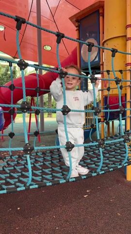 A young boy, approximately two to three years old, is playing on a playground structure. He is wearing a white sweat suit and white tennis shoes. The suit has the phrase "Future goals" printed on the front in cursive. The boy is standing on a blue rope net bridge, part of the play structure. He is holding onto the ropes above him with both hands. He is smiling and appears excited. The playground has a red and yellow color scheme. A red slide is visible in the background. The ground beneath the boy is brown and appears to be a soft, padded surface. There are trees visible in the background, indicating the playground is located in a park-like setting.