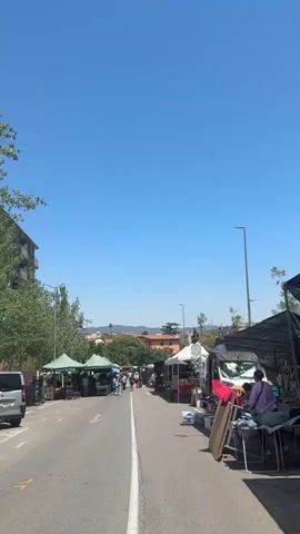 This is a vertical, eye-level, outdoor shot of a street market on a clear, sunny day. The road is paved with asphalt and marked with a white line down the center. On both sides of the street, stalls are set up under various types of awnings, including green, white, and black. The stalls appear to be selling a variety of goods, with items like clothing, household items, and what might be food visible.

On the left side of the street, there are trees and a multi-story building. On the right side, there are more stalls and a white van. Several people are visible browsing the stalls and walking down the street. The overall impression is of a bustling community market. The background shows a slightly hazy view of hills and buildings, indicating a semi-urban or suburban setting. The sky is a clear, bright blue with no visible clouds.