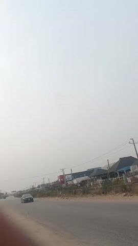 The video shows a road on a hazy day. There are buildings on the side of the road, as well as power lines.  A few cars drive on the road. The shoulder of the road is unpaved.  A glimpse of a person's hand is visible on the left side of the frame.