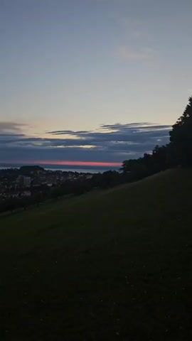 A low-angle, wide shot captures a grassy hill in the foreground, sloping downward towards a distant town nestled beside a body of water. The sky above transitions from a deep blue at the top to lighter shades of pink and orange near the horizon, where a thin band of vibrant red delineates the sunset. Dark clouds are scattered across the sky, adding texture to the scene. The town appears as a collection of buildings, some taller and more modern, interspersed with trees. The overall impression is one of a serene twilight landscape.