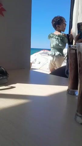 The video shows a young child standing near an opening, looking out at a beach scene. The child is wearing a green, off-the-shoulder top with ruffles and light-colored shorts, along with dark sandals. The child's hair is short and dark. The background reveals a bright blue sky and the ocean. On the beach, there appears to be a white tent or shelter. In the foreground, a wooden table is visible, with two glasses of what looks like chocolate milk or a similar beverage placed on it. Sunlight is casting shadows across the table. The child is actively looking out and gesturing, appearing engaged with the view. The overall impression is a casual outdoor setting, possibly a cafe or restaurant with a view of the sea.