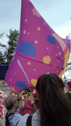 The video shows a crowd of people outdoors, possibly at a festival or event. They are holding up a large, pink inflatable object decorated with blue clouds, yellow stars, and pink flowers. Some children are visible in the crowd, and one is being held by an adult. The background includes trees and a stage with lighting equipment. The overall atmosphere appears festive and active.