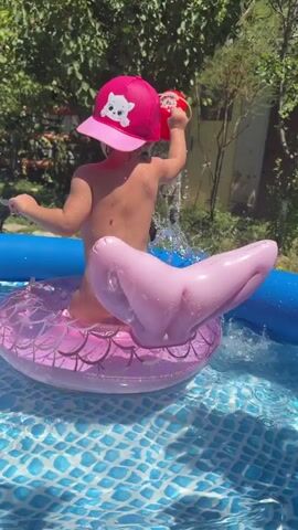 A child wearing a pink hat with a white cartoon cat design is sitting on a pink inflatable ring shaped like a mermaid's tail in a blue inflatable swimming pool. The child is holding a red toy and appears to be playing with water. The pool is set up outdoors in a grassy area with trees and a fence in the background. The water in the pool is clear and reflects the sunlight.