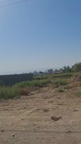 The video shows a dusty, unpaved road in a rural or semi-rural area. The sky is clear and blue. To the left, there is a low, rough brick wall and some overgrown vegetation. Further back, there is a building with a concrete foundation and what appears to be a shipping container. In the center, there is more dense, green vegetation. To the right, the road continues, and beyond that, there is more vegetation and some distant structures that could be buildings or unfinished construction. The video pans slowly to the right, revealing more of the landscape. There are no people or animals visible in the frame. The overall impression is of an undeveloped or developing area with signs of construction.