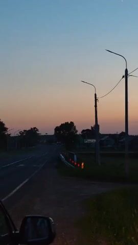 A stork stands silhouetted on top of a lamppost against a dusky sky. In the background, a second lamppost and power lines are visible, along with the dark shapes of trees and buildings. The sky transitions from a pale blue at the top to a soft orange and pink near the horizon, indicating either sunrise or sunset. A small, bright star is visible in the upper portion of the sky. The scene appears to be near a road, suggested by a guardrail and a road sign visible in the lower left corner.