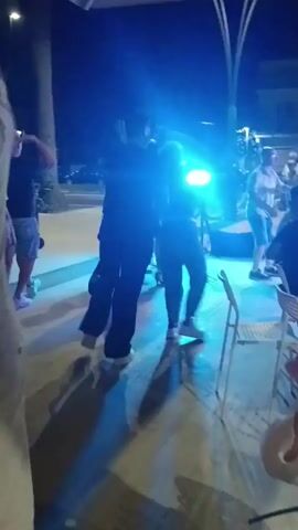 A group of people are dancing in what appears to be an outdoor venue at night. The lighting is dim with accents of purple and pink. In the foreground, a woman in a black top and tan pants is dancing with her mouth open. To her right, a man in a light-colored polo shirt and tan pants is dancing. In the background, several other couples are dancing, and some people are standing and watching. The ground is paved, and there are some lights and structures visible in the background, suggesting an outdoor patio or plaza.