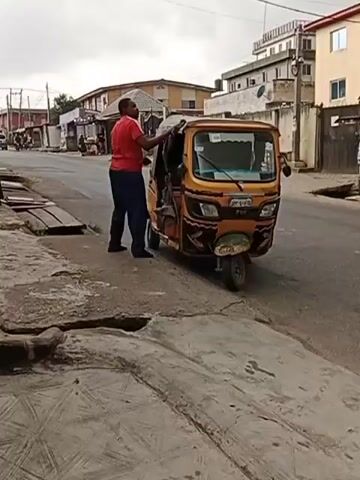 A man wearing a red shirt and dark pants is standing next to a yellow auto rickshaw. He's on the passenger side, near the front. The rickshaw is parked on the side of a street in a residential area. The man appears to be interacting with someone inside the rickshaw. The road is paved but shows signs of wear and tear, and there's a sidewalk-like area next to the road that's also damaged. Other buildings and people are visible in the background. The weather appears overcast.