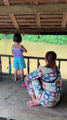 A woman in pajamas with Snoopy characters sits on a wooden floor, facing a muddy river. A child stands next to her, looking at the river. The child wears a purple and white top and light blue shorts. They are on a raised platform with a wooden railing adorned with colorful ribbons. The river is murky brown, and lush green trees line the opposite bank. The ceiling above them is made of wooden planks, and string lights hang from it.