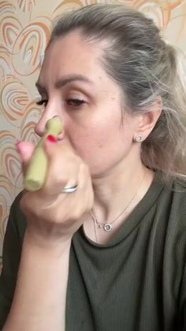 A woman with blonde hair pulled back and wearing a green shirt is holding a small, light green cylindrical device with a silver rollerball tip to the skin under her right eye. She is looking down and to her left as she moves the device across her under-eye area. Her left hand, adorned with a ring, is holding the device. The background is a wall with a patterned wallpaper in shades of beige, brown, and orange.