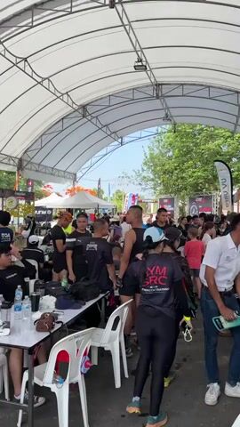 The video captures a bustling scene under a white arched tent structure, filled with people, tables, and banners, suggesting a public event or gathering. The crowd is composed of individuals of varying ages and ethnicities, dressed in casual clothing, athletic gear, and uniforms, indicating a mix of participants, organizers, and spectators. Several people are standing around tables with water bottles and personal items, while others are moving through the space, conversing, or examining displays. The environment features event branding on flags and signs, with trees and blue sky visible in the background, further establishing the outdoor setting.