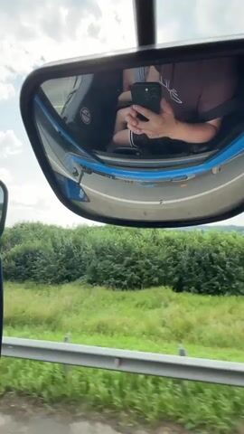 The video shows a view from inside a vehicle. The camera is positioned to capture the side-view mirror, which reflects a person holding a smartphone and taking a selfie. Outside the vehicle, there is a green landscape with trees and fields under a cloudy sky. A river is visible in the distance. The vehicle appears to be moving, as indicated by the motion blur in the landscape.