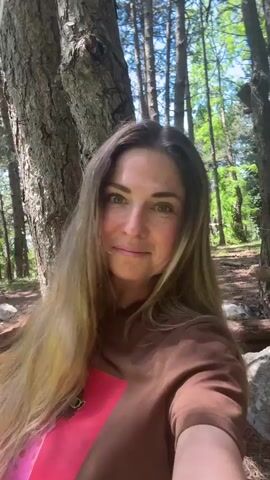 A woman is filming herself in a forest setting. She is wearing a brown t-shirt with a pink graphic on the front. The woman has long blonde hair and is looking at the camera. She is standing in a wooded area with tall trees. The sun is shining through the trees, creating bright light. The woman appears to be smiling. She then looks up at the sky and closes her eyes. She is holding onto a rope which is attached to a tree. The camera then tilts upwards, showing the sun and the trees.