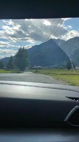 The video shows a view from inside a car. The camera is looking out through the windshield. The car is driving on a paved road, and the landscape outside is mountainous and rural. There are trees, fields, and some buildings visible in the distance. The sky is cloudy but with some patches of blue. The dashboard of the car is visible in the foreground. The video pans slightly to the right as the car moves.
