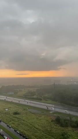 A drone shot captures a wide view of a rain-swept landscape under a cloudy, orange-hued sky. In the foreground, a patch of lush green grass is dotted with parked cars. A wet, multi-lane highway stretches across the middle ground, with several cars driving on it. Beyond the highway, a vast expanse of green fields and dense trees stretches to the horizon. In the far distance, several buildings and a tall tower are visible through the mist and rain. The sky is filled with dark, heavy clouds, with a bright band of orange light peeking through the horizon, suggesting either a sunset or sunrise. The overall impression is one of a dramatic, atmospheric weather event.