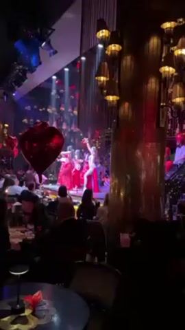 The video captures a performance in an opulent nightclub. Three belly dancers, dressed in red and silver sequined costumes, perform on a lit stage for an audience seated at tables. A large, red heart-shaped balloon is positioned near the stage. The interior is decorated with intricate gold accents, including decorative lighting fixtures and architectural details. The audience is dressed formally and appears to be enjoying the show.