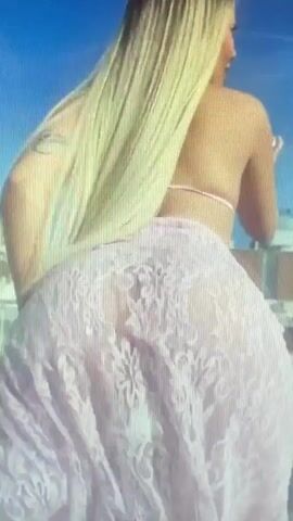 A woman with long blonde hair is viewed from behind. She is wearing a light pink lace garment that appears to be a skirt or dress, and a thin strap visible around her waist. The background shows a blue sky and some buildings.