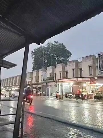 The video shows a street in what appears to be an urban area during rainfall. The shot is taken from beneath a corrugated metal roof, possibly a shelter or awning.  Across the street, there's a row of buildings with a uniform architectural style. Some of the storefronts are open and illuminated. Traffic is light but visible, with cars and motorcycles moving along the wet road. The sky is overcast, contributing to the overall gray and damp atmosphere. The focus is sharp, showing the details of the scene clearly despite the rainy weather.