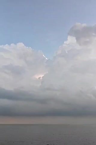 The video shows a view of the ocean beneath a cloudy sky. The clouds are predominantly white and grey, forming large, billowy shapes that dominate the upper portion of the frame. There is a small opening of blue sky visible between the cloud formations. The ocean is a dark grey color and calm with a small boat in the distance.