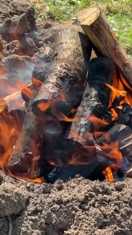A close-up shot shows logs burning in a campfire. The fire is intense with bright orange flames and wisps of grey smoke. The logs are stacked in a tepee formation, with some pieces appearing charred and others still showing bark. The fire is built on a mound of dirt, and in the background, there is green grass, suggesting an outdoor setting during the daytime. There is no visible text in the image. The scene is focused on the fire itself, conveying a sense of warmth and natural energy. The location context provided, "Радашковіцкі сельскі Савет, Belarus," places the scene within a rural area of Belarus. The image is devoid of any people, focusing solely on the natural elements of the campfire. The lighting suggests it is daytime, with the sun illuminating the scene and the grass in the background. The overall mood is one of natural beauty and a peaceful outdoor experience.