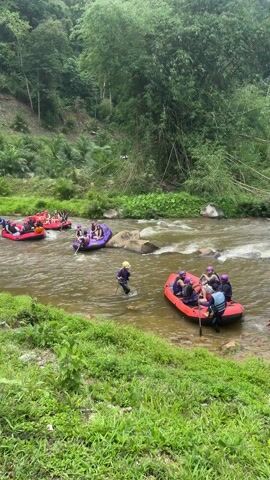Several inflatable rafts with people are navigating a river through a forested area. The rafts are different colors, including red, purple, and blue. Most of the people are wearing life vests and helmets. Some people are using paddles to steer the rafts. One person is standing in the water with a long pole, guiding one of the rafts. The river flows through a green, lush environment with trees and vegetation lining the banks. The water is slightly murky and there are some rocks visible in the riverbed. The camera angle is from an elevated position on the riverbank, showing a wider view of the activity.