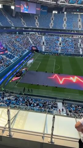 The video shows a wide shot of a football stadium filled with spectators. The field is green with white markings. A large banner or tarp with a red and yellow lightning bolt design is spread across the field. Several people are visible on the field, some holding the banner. In the stands, the crowd is predominantly wearing blue and white. A large screen at the top of the stadium displays two figures. The overall atmosphere appears to be during a pre-game event or ceremony. The viewpoint is from a higher tier of seating, looking down at the field and the opposite side of the stadium. The lighting suggests it is daytime with natural light entering the stadium.