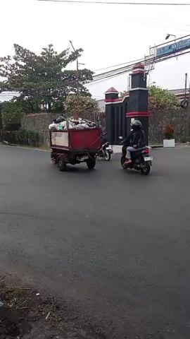 A collection of still frames shows a street scene in what appears to be a city or town. The road surface is wet, suggesting recent rain. On the left side of the road, a three-wheeled vehicle, possibly a garbage truck, is visible. To its right, a person on a scooter is traveling. Further down the road, more scooters are seen, some carrying multiple passengers. In the background, there is a large green tree and a distinctive black and red gate or structure. The overall atmosphere seems to be one of everyday traffic and activity on a cloudy day.