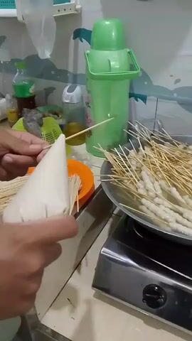 A person is using a piping bag to add batter to a skewer of raw meatballs. The skewer is held over an orange plate filled with more empty skewers. A large pan filled with skewers of meatballs is visible in the background, sitting on a stovetop. A green thermos and a green bottle are on a shelf behind the pan.