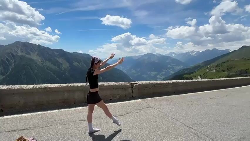 A young woman with dark hair and a headscarf is performing a handstand on an asphalt road. She wears a black crop top, black shorts, white socks, and white shoes. Her body is inverted, with her hands on the ground and her legs extended upwards, forming an 'X' shape. The background features a breathtaking mountainous landscape with lush green slopes and a valley under a bright blue sky dotted with white clouds. A concrete barrier runs along the side of the road. The woman transitions from a forward-leaning pose to the handstand and then back to the standing pose.