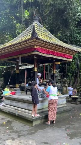 A group of people are gathered around a tiered, temple-like structure in a lush, green environment. The structure has a yellow, tiled roof adorned with red fabric and gold trim. A man wearing a white outfit and a white cap sits on a raised platform within the structure, interacting with two females. One female, wearing a black top and shorts, stands to the left of the platform. The other female, wearing a white top and a patterned skirt, stands to the right. In the background, a lush green cliff face features numerous small statues and decorative umbrellas, with water cascading down from some of the statues. Two people, a male and a female, are seated in meditation poses on yoga mats in front of a long, draped table that displays offerings. The ground around the structure is wet and scattered with colorful flower petals.