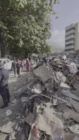 The video shows the aftermath of a disaster, with a large pile of rubble in the foreground. The rubble consists of concrete, metal, and other debris, indicating the collapse of a building. In the background, a few people are visible walking and observing the destruction. Some are wearing hard hats, suggesting they might be part of a rescue or recovery effort. A large building with multiple stories stands partially intact, but also shows signs of damage and appears to be leaning. The sky is bright and sunny, contrasting with the scene of devastation. Trees line the street, and power lines are visible overhead. The presence of scattered personal items and appliances like a refrigerator suggests that the building was likely residential or commercial. The overall scene conveys a sense of destruction and the need for cleanup and recovery.