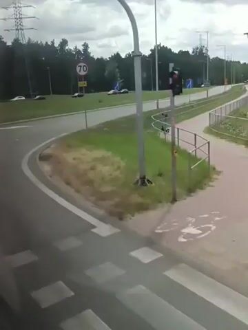The video shows a view from a vehicle traveling on a road. The road curves to the right. On the left side of the road, there is a grassy area with several cars parked. In the distance, there are trees and a large electrical tower. A speed limit sign indicating 70 is visible. There are also road signs with arrows indicating directions. The sky is overcast. The video appears to be a recording from a dash camera or a similar device.