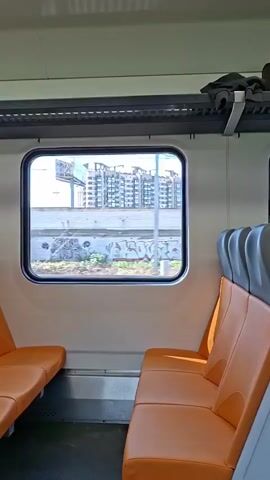 The video shows the interior of a train carriage. The seats are orange and grey. Through the window, buildings and some greenery can be seen. The train appears to be moving, as the scenery outside is blurred. There are no people clearly visible in the frame, though there may be blurred figures outside the train.