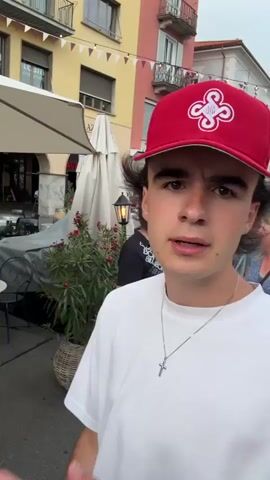 The video shows a young man wearing a red baseball cap and a white t-shirt. He is standing outdoors in what appears to be a town square or street. In the background, there are buildings with colorful facades, umbrellas from outdoor seating areas of restaurants, and string lights overhead. There are other people in the background, some walking and some seated at tables. The young man makes several hand gestures, including a rock and roll sign, and points at the camera. He also appears to be speaking, but the audio is not transcribed. The overall atmosphere seems to be that of a lively outdoor event or gathering.