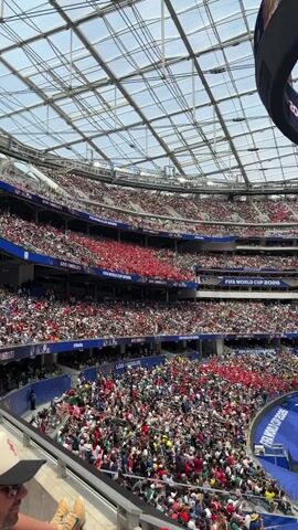 The video shows a wide shot of a soccer stadium filled with a large crowd. The stadium has multiple levels of seating, with many spectators wearing red and white. The roof of the stadium is visible, constructed with a grid of metal beams and translucent panels. There are large screens displaying the score of a match, which reads "2:0" with team logos. The field itself is a vibrant green soccer pitch with players in white and red uniforms scattered across it. Banners and signs indicating "FIFA WORLD CUP 2026" and "LOS ANGELES" are visible throughout the stadium, suggesting the event is a World Cup qualifier or related event in Los Angeles. The overall atmosphere appears to be one of excitement and anticipation.