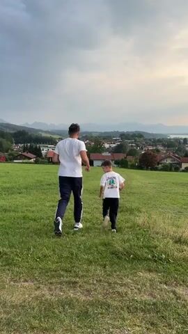 Two people, an adult and a child, run away from the camera across a grassy field. The adult wears a white t-shirt and dark pants, while the child wears a white t-shirt and dark pants. Behind them, a village with houses and trees is nestled in a valley, with mountains in the distance under a cloudy sky.