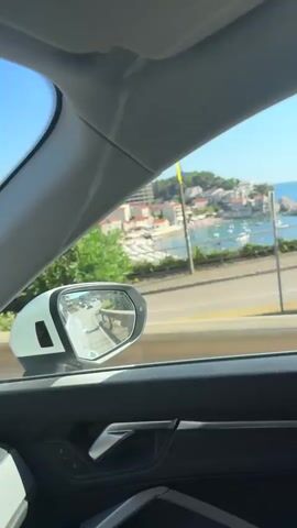 The video is shot from inside a car looking out the passenger window. The camera pans slightly, showing a side view mirror reflecting the road behind. The car appears to be moving along a coastal road, with lush green foliage and pink flowers visible outside. In the distance, there are buildings and the sea. The interior of the car, including the door panel and window frame, is also visible. The sky is clear and blue. The video has no sound.