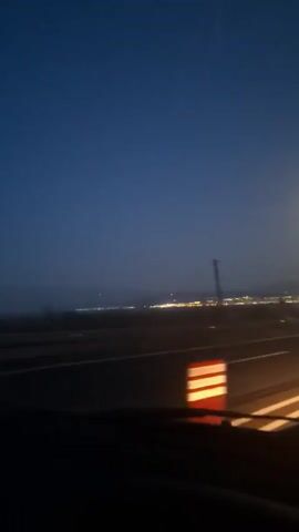 The video shows a night-time view from inside a moving vehicle on a highway. The sky is a deep blue, and streetlights and building lights are visible in the distance. Red and white striped traffic cones are spaced along the roadside. There are white lines marking the lanes on the road.